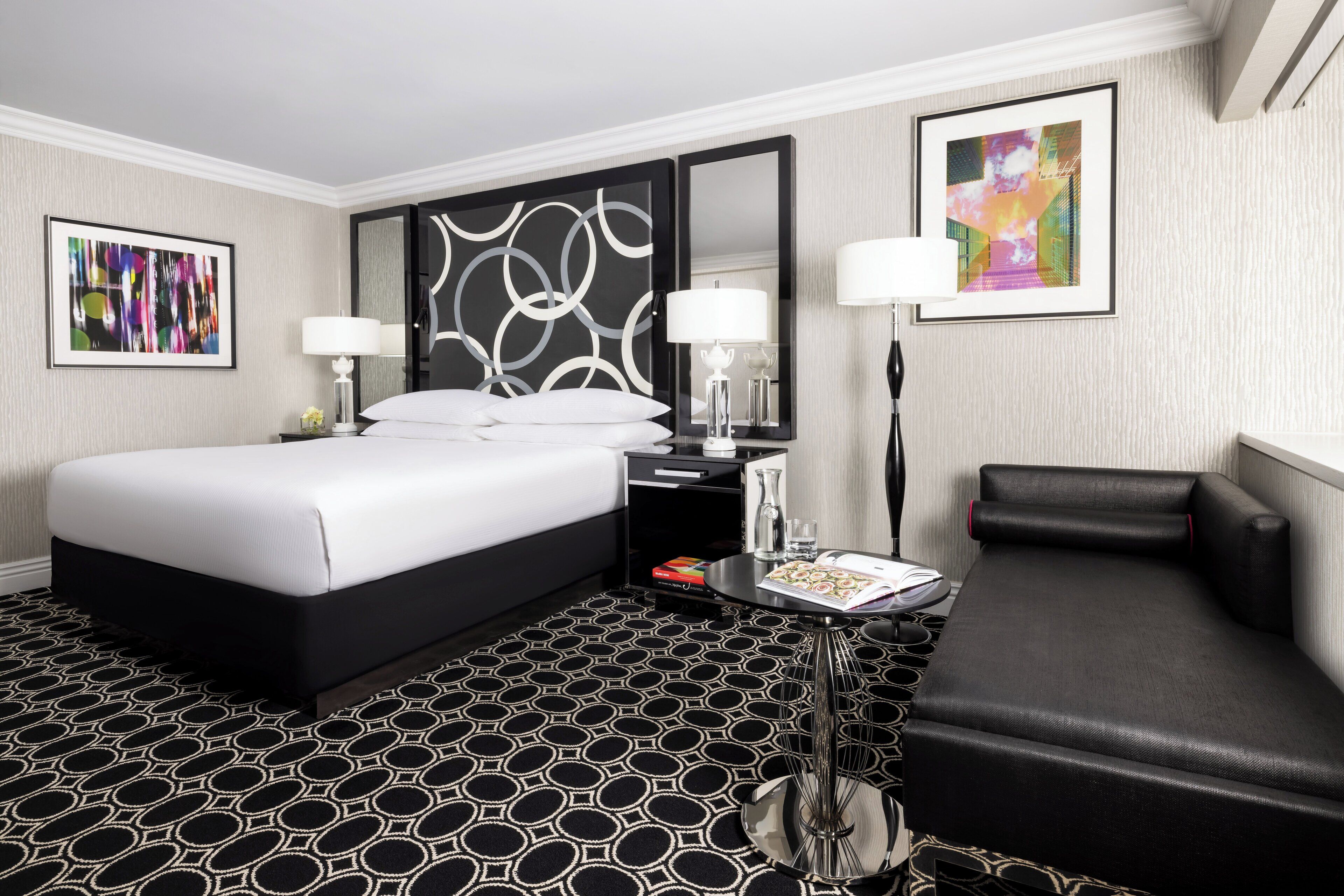 A hotel room with a large bed, a black leather sofa, a small table with a lamp, and a mirror.