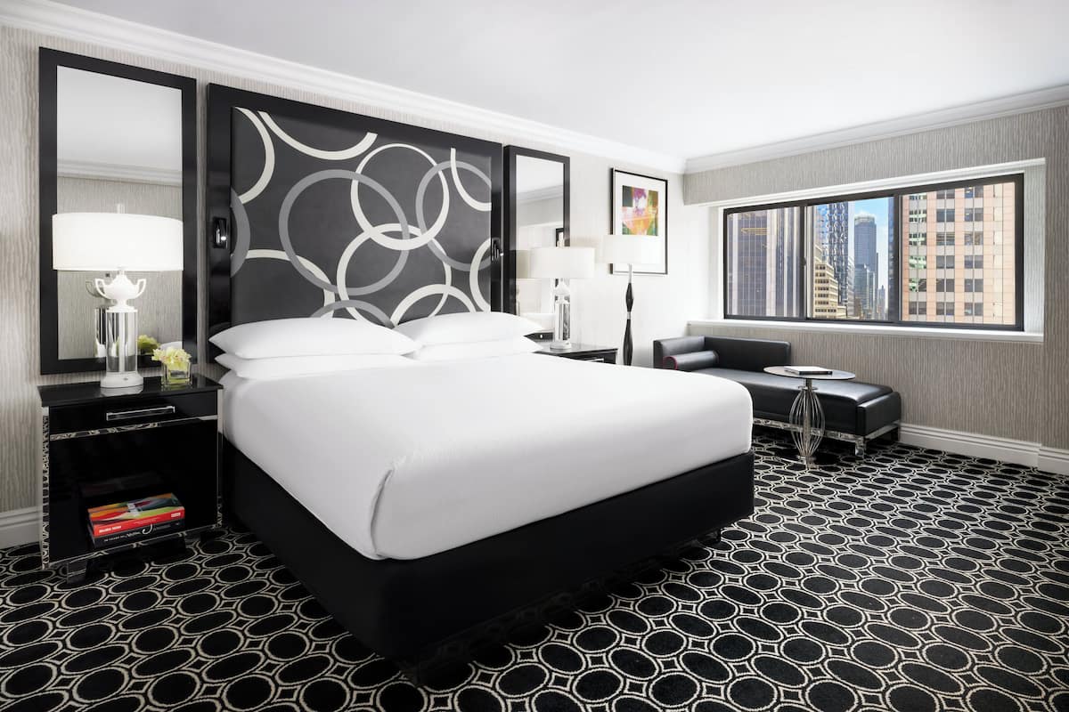 Et moderne hotelværelse med en stor seng, et sengebord med lampe, et lille bord med lampe og udsigt over byen gennem et stort vindue.