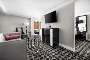 A modern hotel room with a grey sofa, a black coffee table, a flat-screen TV mounted on the wall, and a patterned floor.
