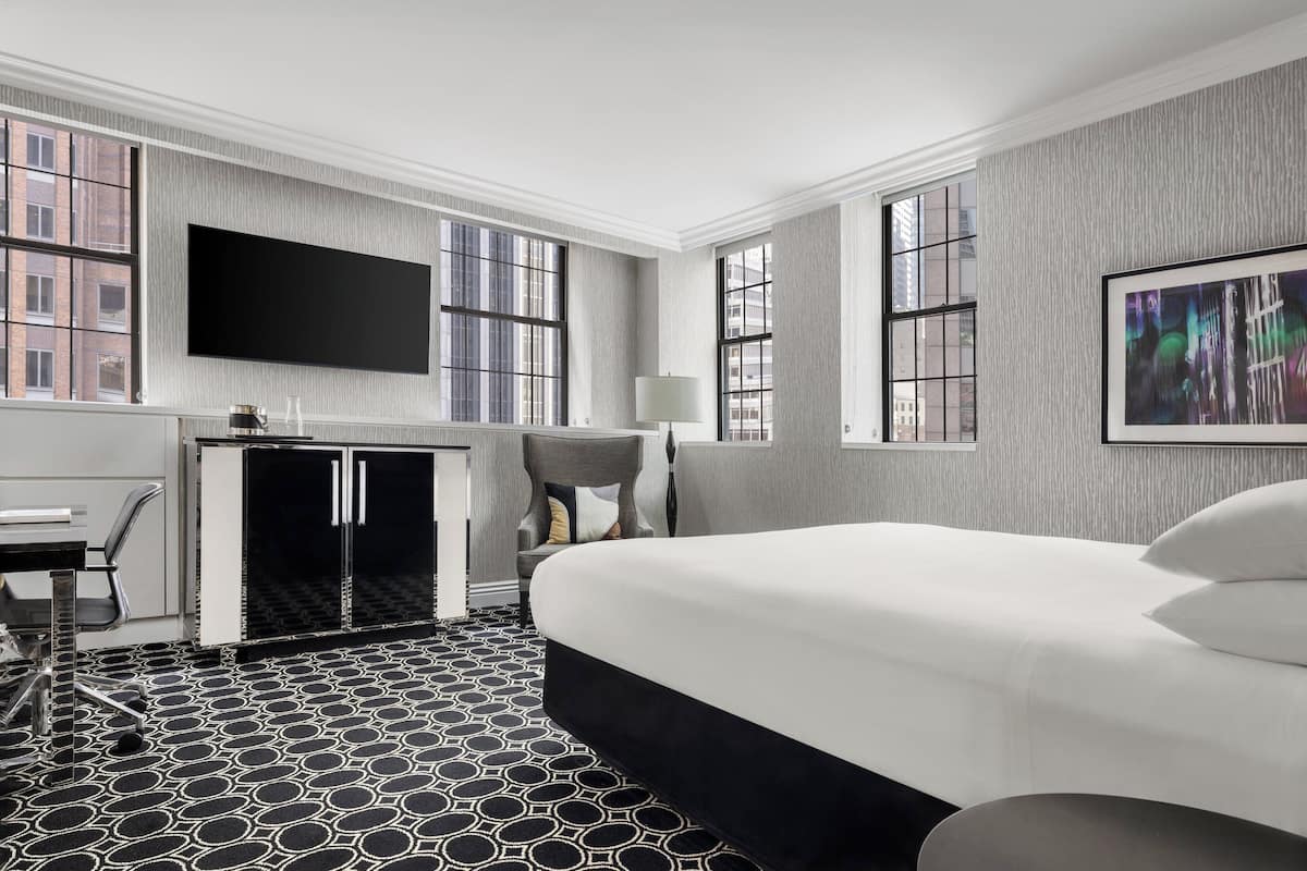 A hotel room with a large bed, a desk with a chair, a television mounted on the wall, and a window with a city view.