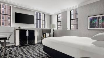 A hotel room with a large bed, a desk with a chair, a television mounted on the wall, and a window with a city view.