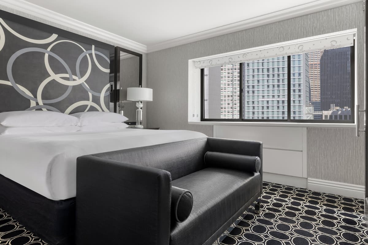 A modern hotel room with a large bed, a sofa, a nightstand, a lamp, and a window with a city view.