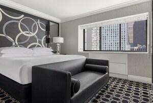 Quarto de hotel moderno com uma cama grande, um sofá, uma mesa de cabeceira, um abajur e uma janela com vista para a cidade.