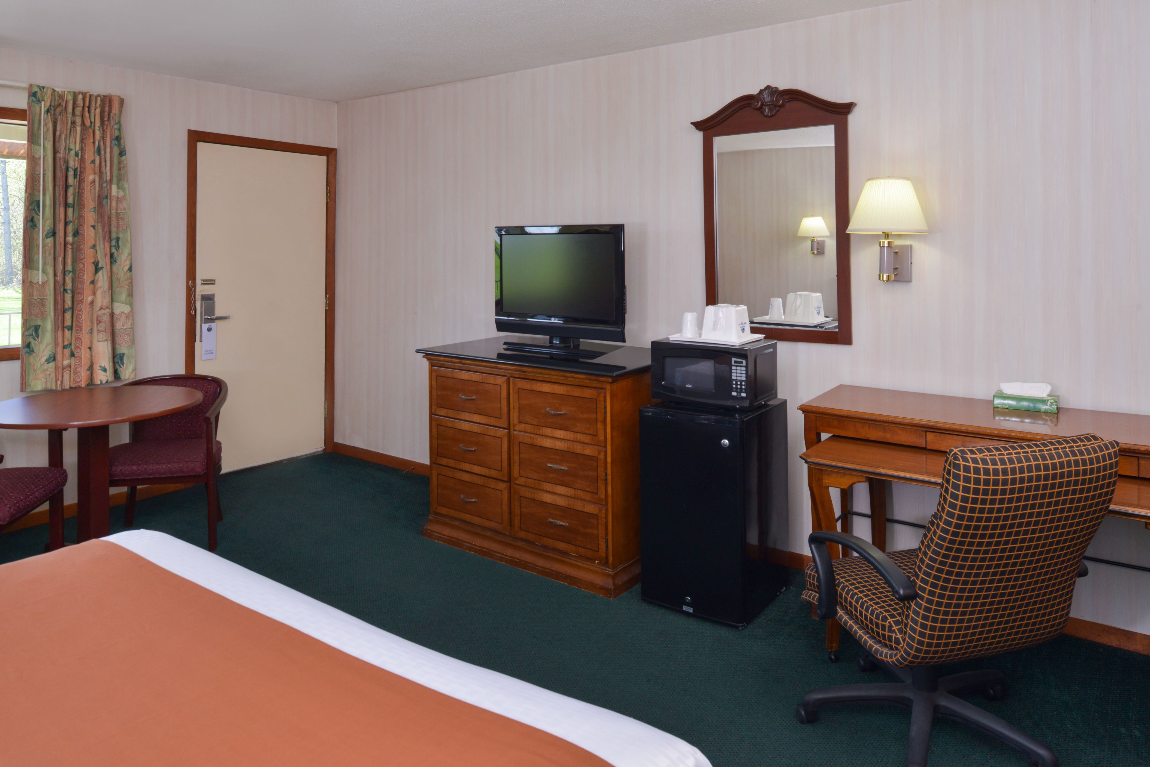 A hotel room with a bed, a desk with a computer, a television, a microwave, a chair, and a dresser.