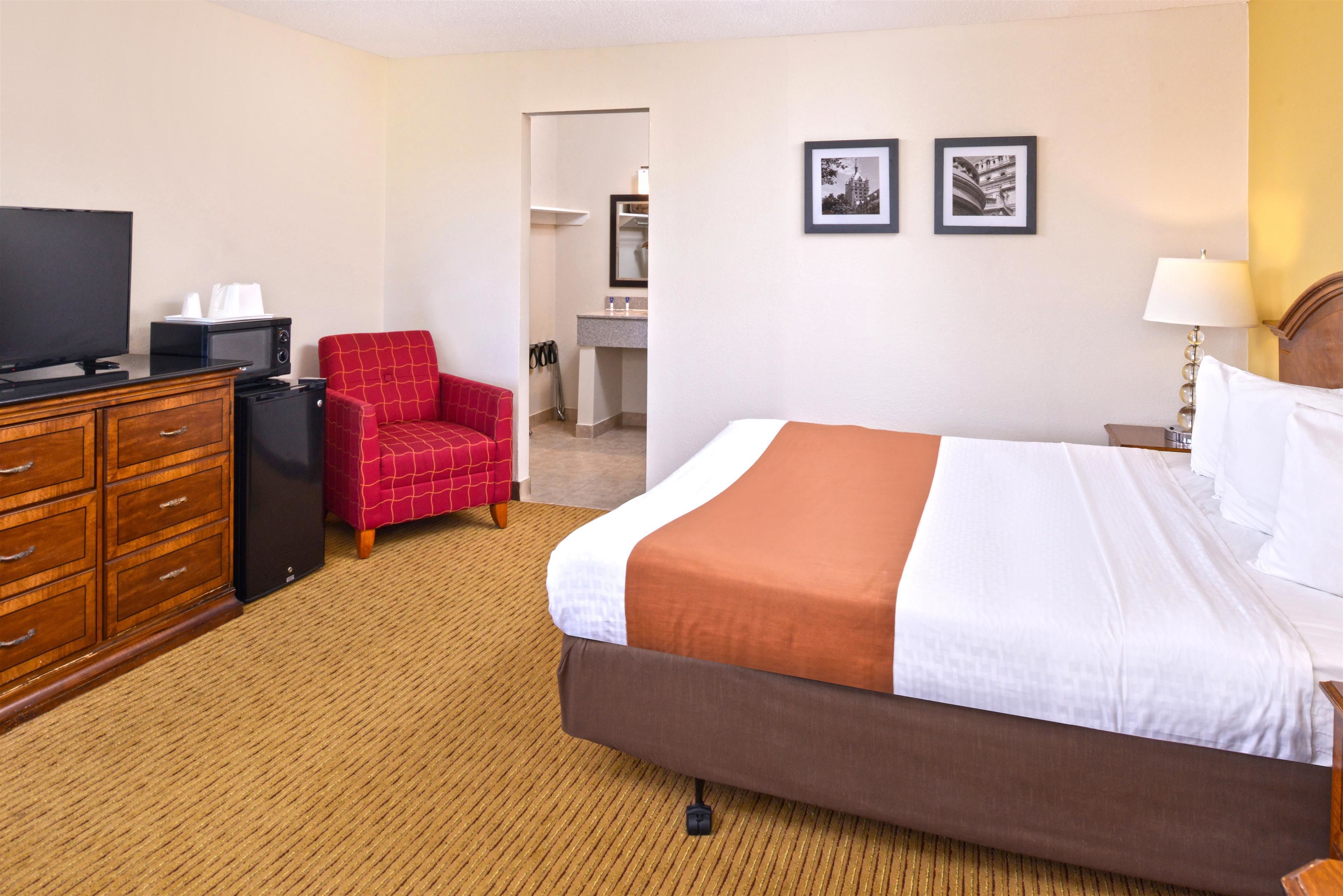 A hotel room with a bed, a TV, a red chair, a desk, and a bathroom visible through an open door.