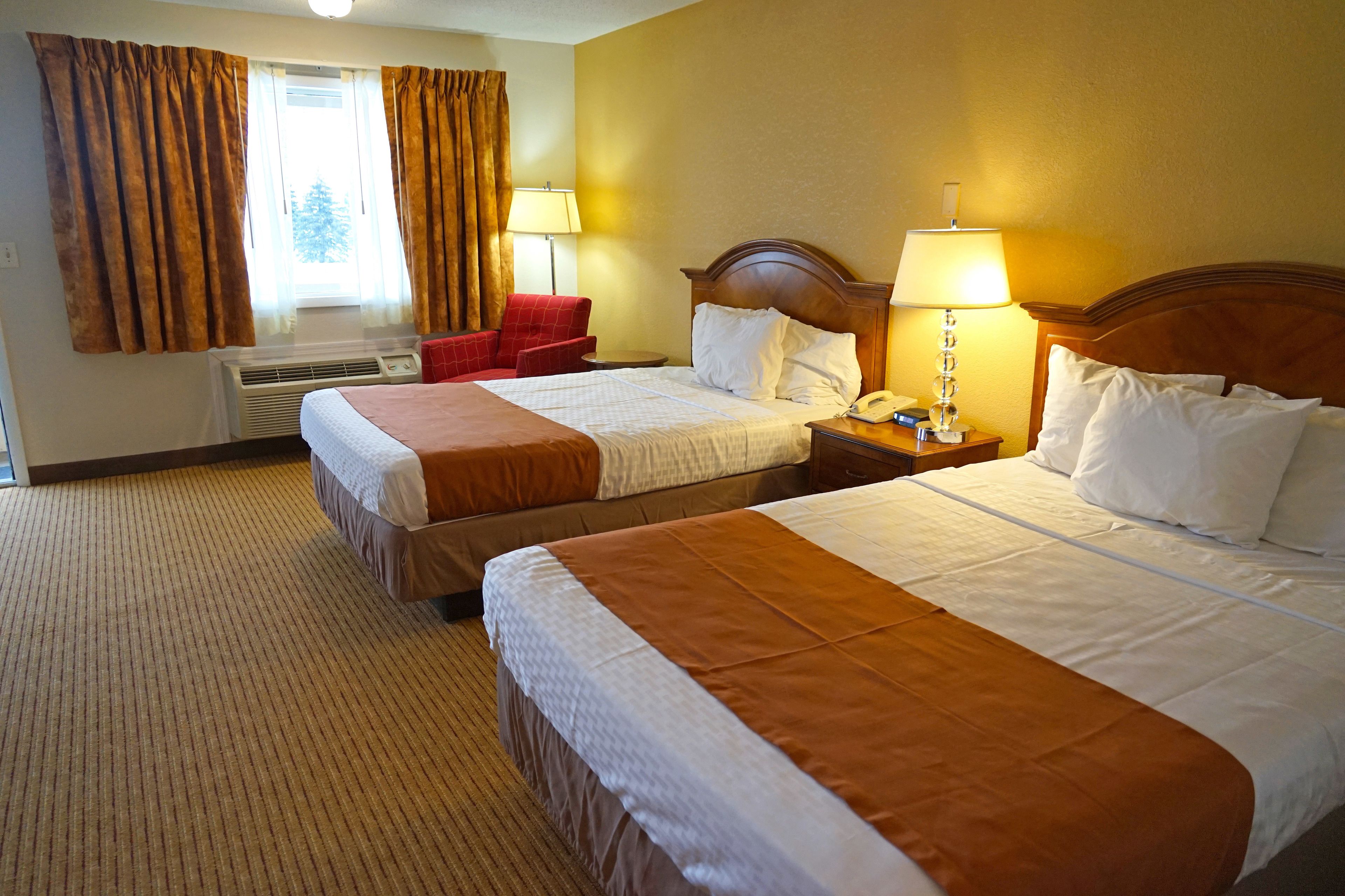 A hotel room with two beds, a red chair, a nightstand with a lamp, and a window with curtains.
