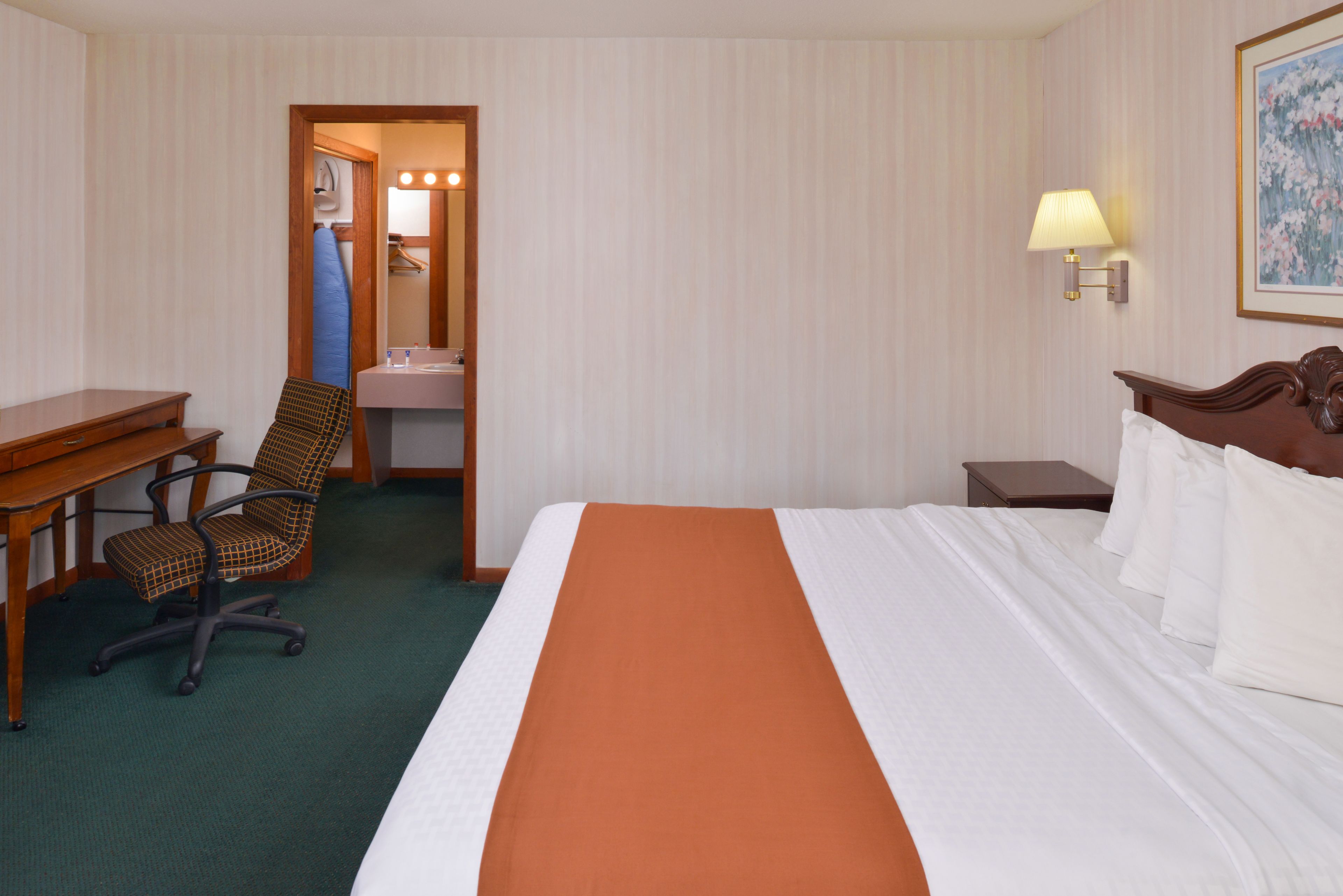 A hotel room with a bed, a desk, a chair, a nightstand, a lamp, and a painting on the wall.