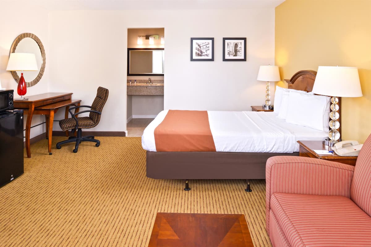 A hotel room with a bed, a desk with a chair, a sofa, a small table with a lamp, a telephone, and a mirror.