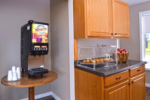 A coffee machine with a display, a small table with cups, and a counter with a fruit bowl and pastries.