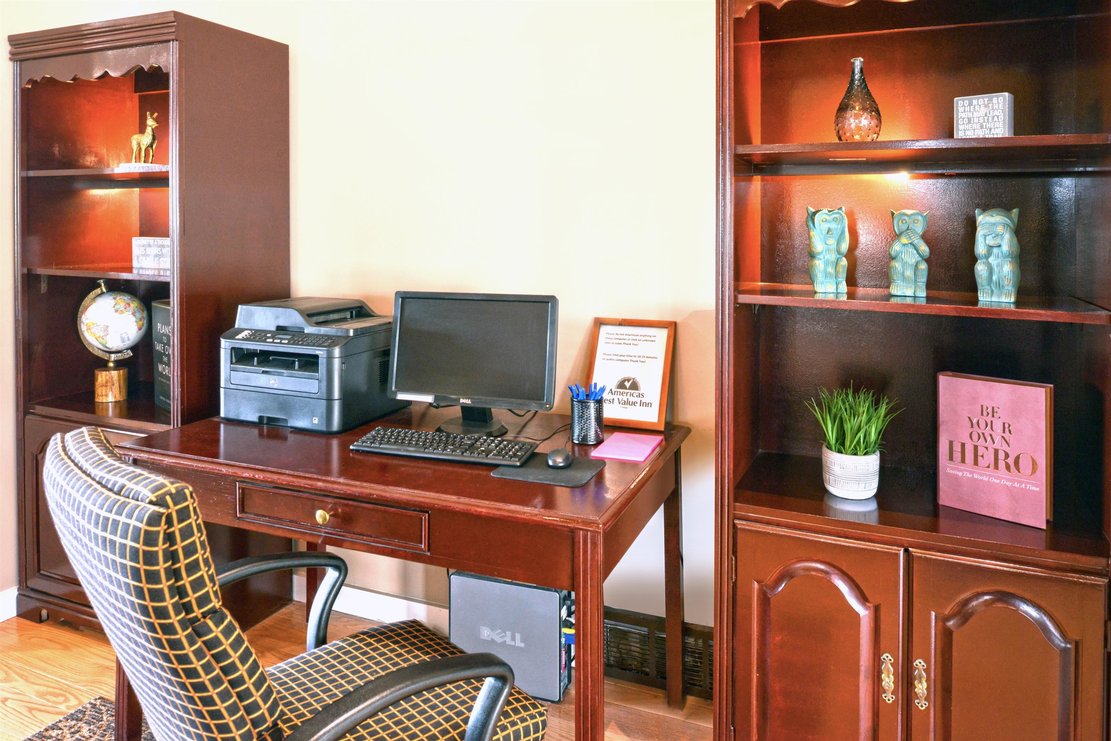 Un escritorio de madera con una computadora, una impresora y un teclado. Hay una silla con un cojín estampado. Se observa una estantería con objetos decorativos y un certificado enmarcado.