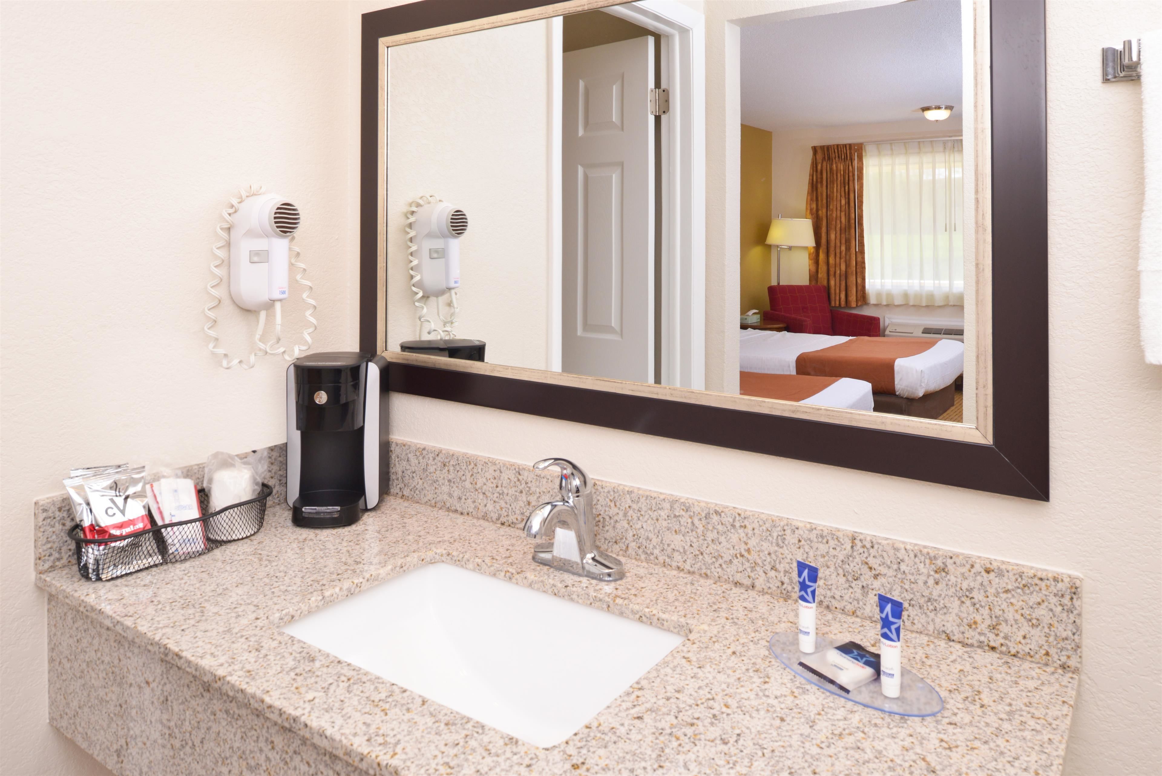 Un baño de hotel con encimera de granito, un lavabo, un espejo, un secador de pelo, una cafetera y artículos de aseo.