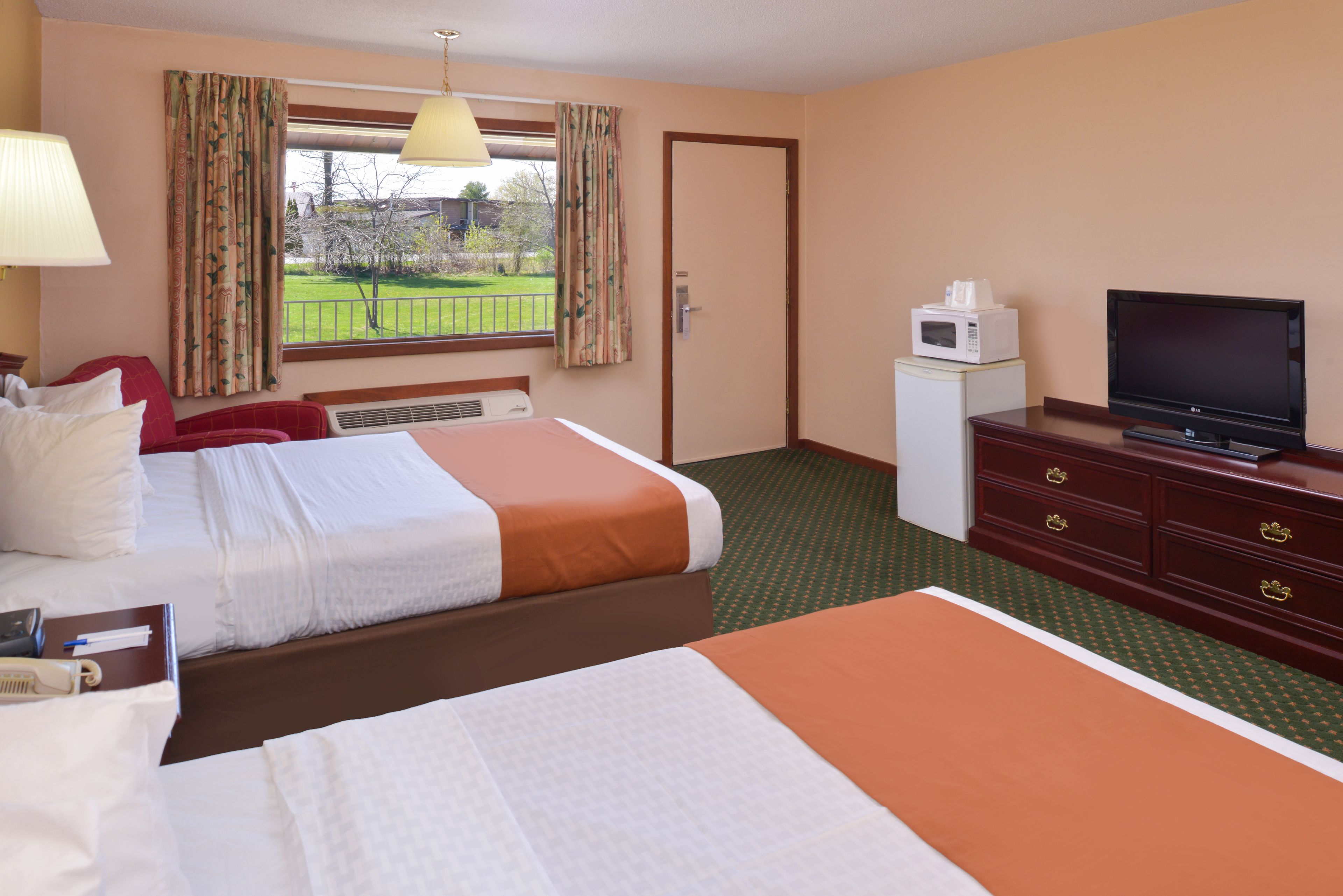 A hotel room with two beds, a television, a microwave, and a view of a green lawn.