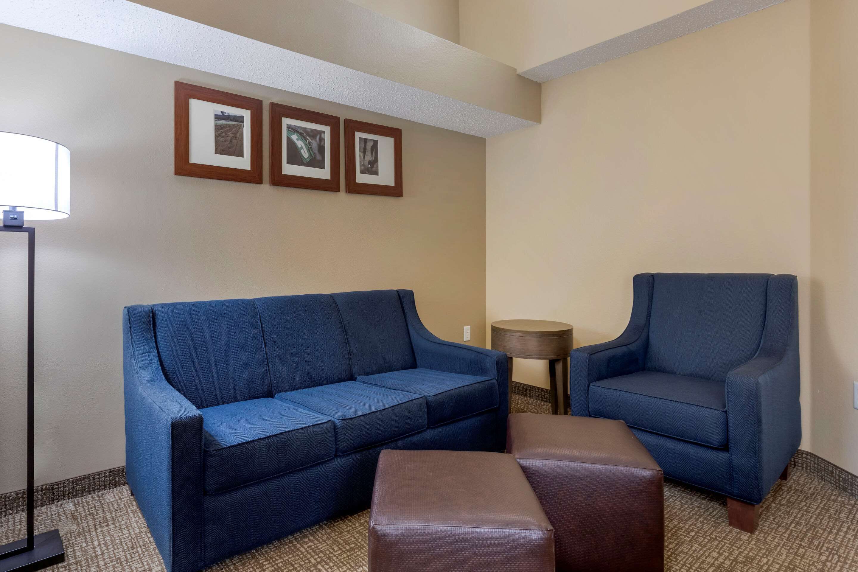 A hotel room with a blue sofa, a brown ottoman, and a blue armchair. There are three framed pictures on the wall and a small round table.