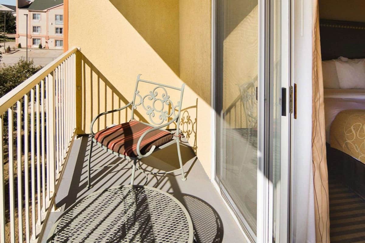 A balcony with a metal chair and a table, a sliding glass door leading to a bedroom with a bed and a pillow with text.