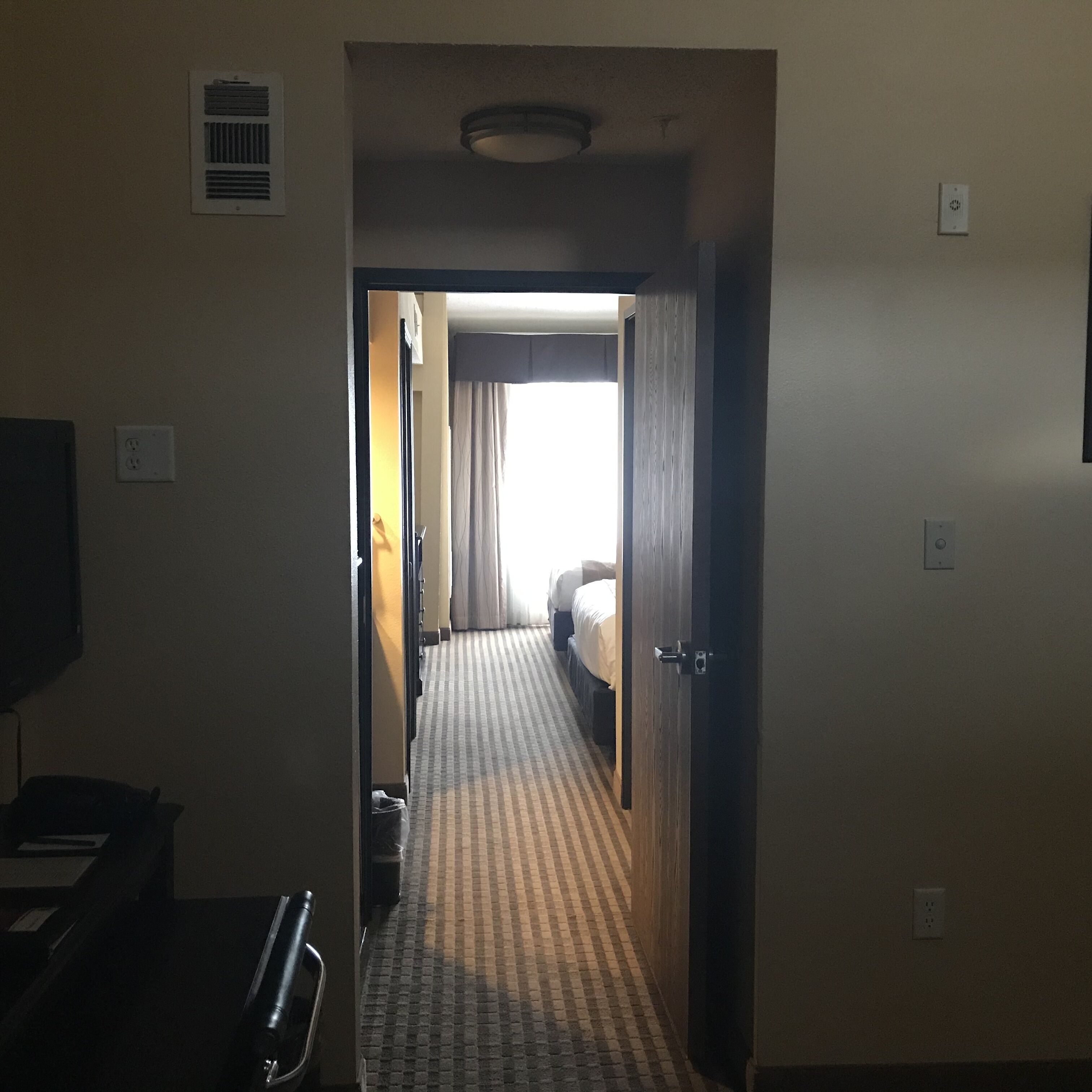 A hotel hallway with a checkered carpet, a bed at the end, and a television on the left.