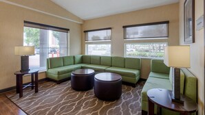 Quarto de hotel com sofá modular verde, dois pufes redondos, uma mesinha de centro com abajur e janelas amplas com persianas.