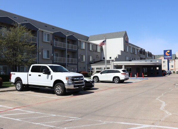 Un estacionamiento con una camioneta pickup blanca y una camioneta SUV blanca estacionadas frente a un hotel con un asta de bandera y un letrero de Comfort Suites.