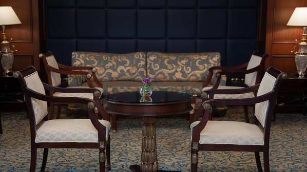 Una habitación con un sofá tapizado en azul oscuro, dos sillas de madera con asientos acolchados, una mesita redonda con un florero y dos lámparas a cada lado del sofá.