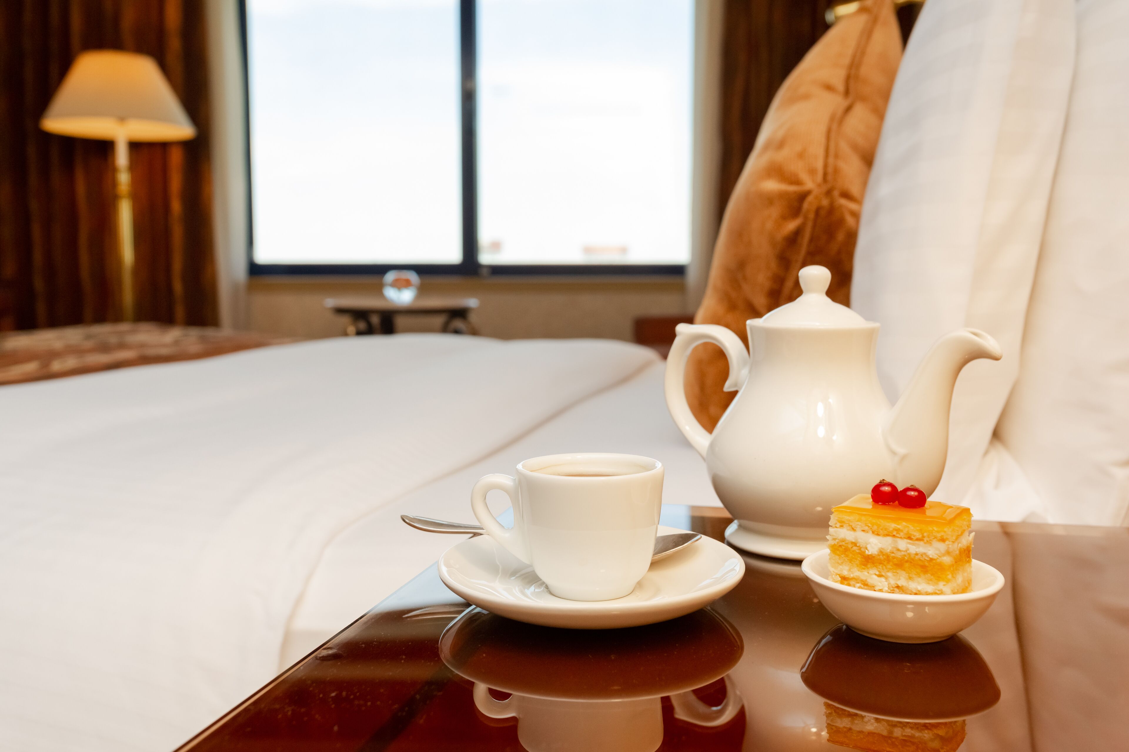 Una cama bien hecha con sábanas blancas, una mesita de noche con una taza de café y un pastel pequeño, y una ventana con cortinas al fondo.