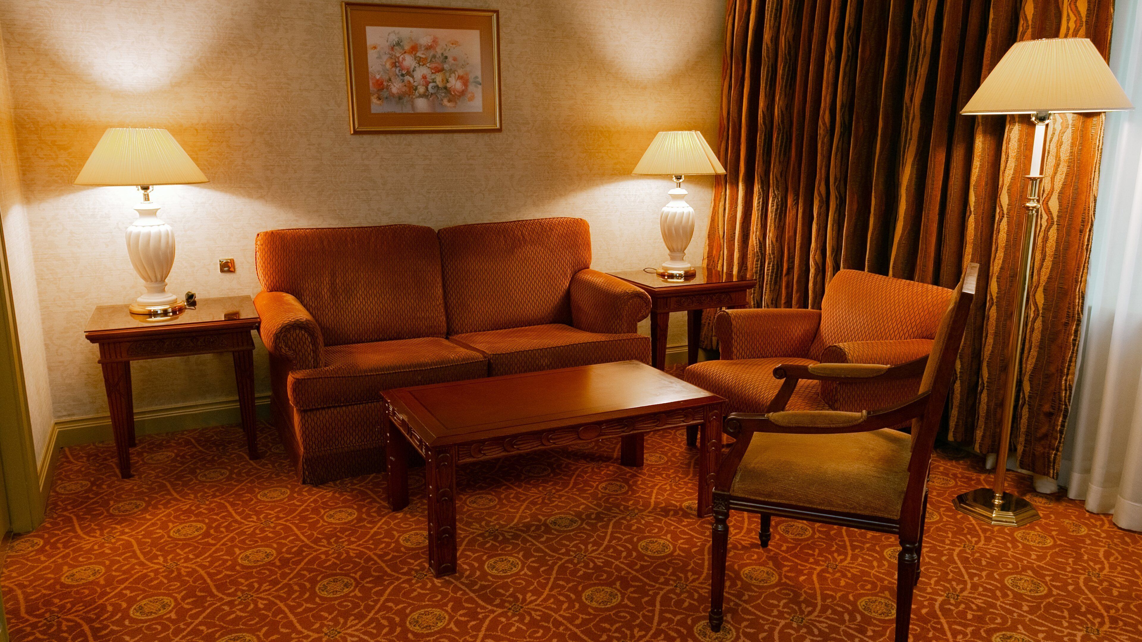 Habitación de hotel con sofá, una silla, una mesa de centro, dos lámparas, un cuadro en la pared y alfombra estampada.