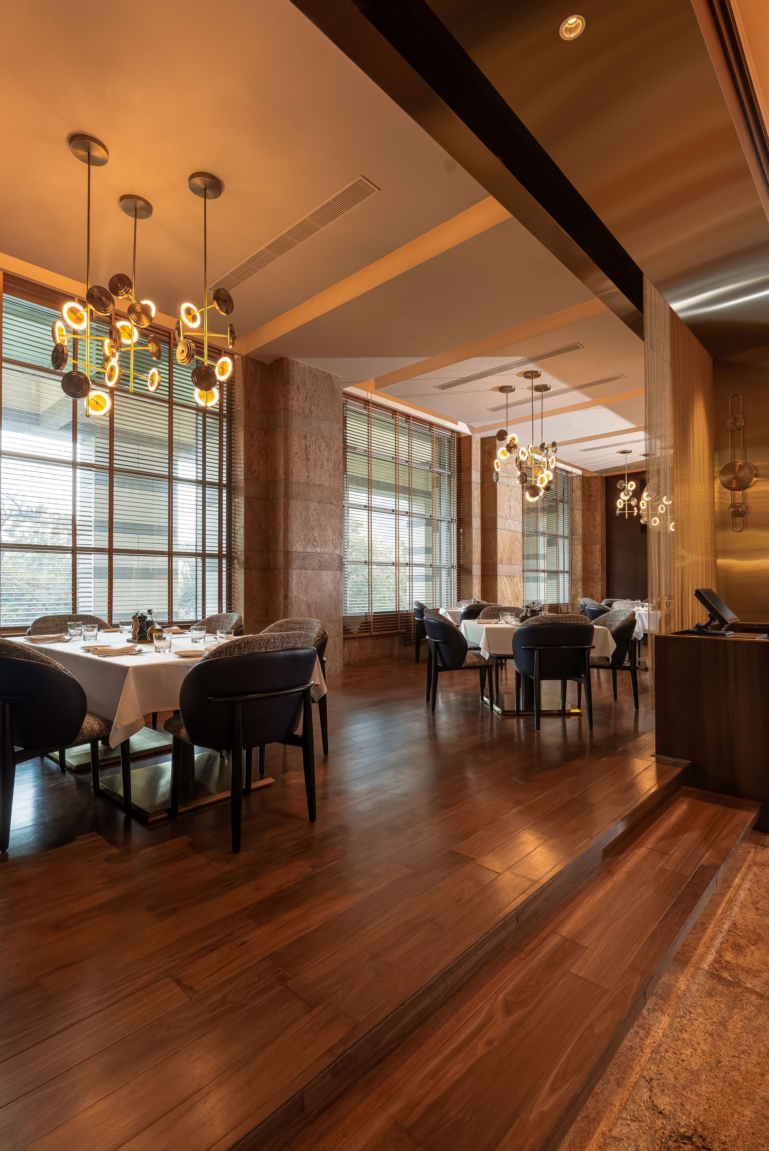 A spacious dining area with multiple tables and chairs, large windows with blinds, and modern lighting fixtures.