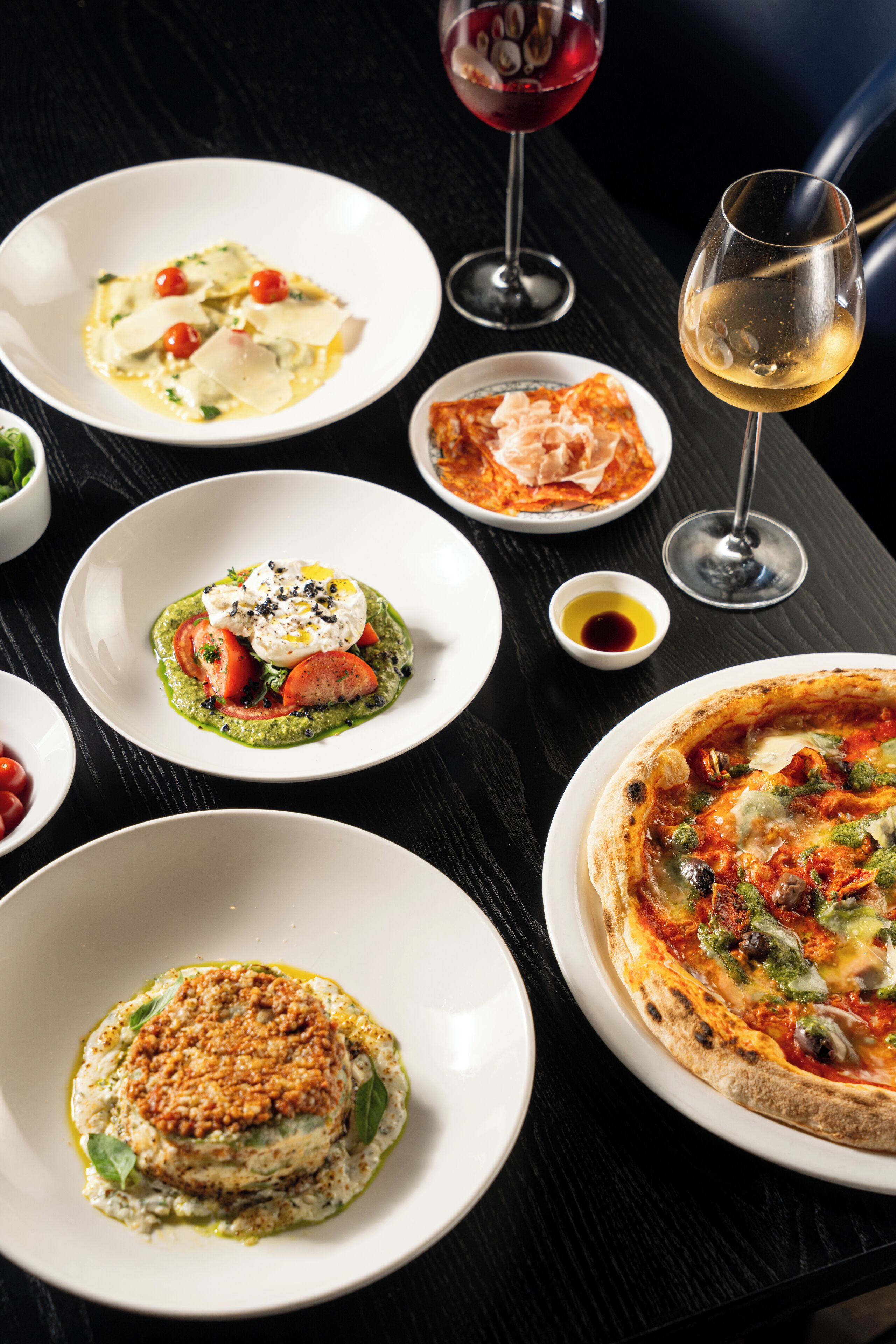 A variety of dishes on a table, including a pizza, pasta, and a salad, with glasses of wine.