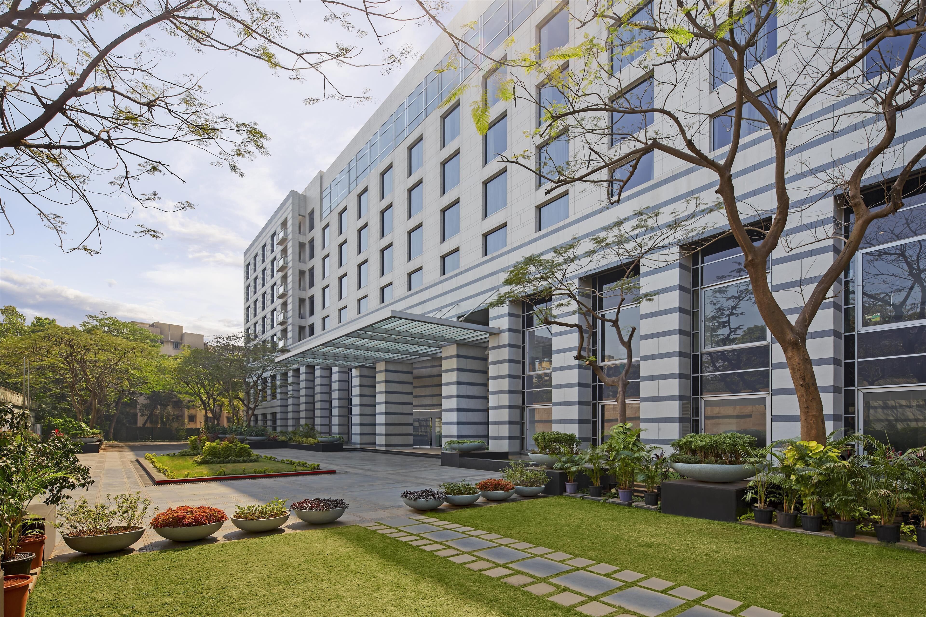 A modern hotel with a glass facade, a covered entrance, and a well-maintained garden with various plants and a pathway.