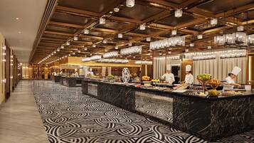 A hotel lobby with a long marble-patterned floor, a high ceiling with hanging lights, and a buffet area with chefs preparing food.