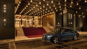 Entrada moderna d'un hotel amb catifes vermelles, un cotxe Mercedes-Benz a l'exterior i un rètol que diu "The Grand Showroom".