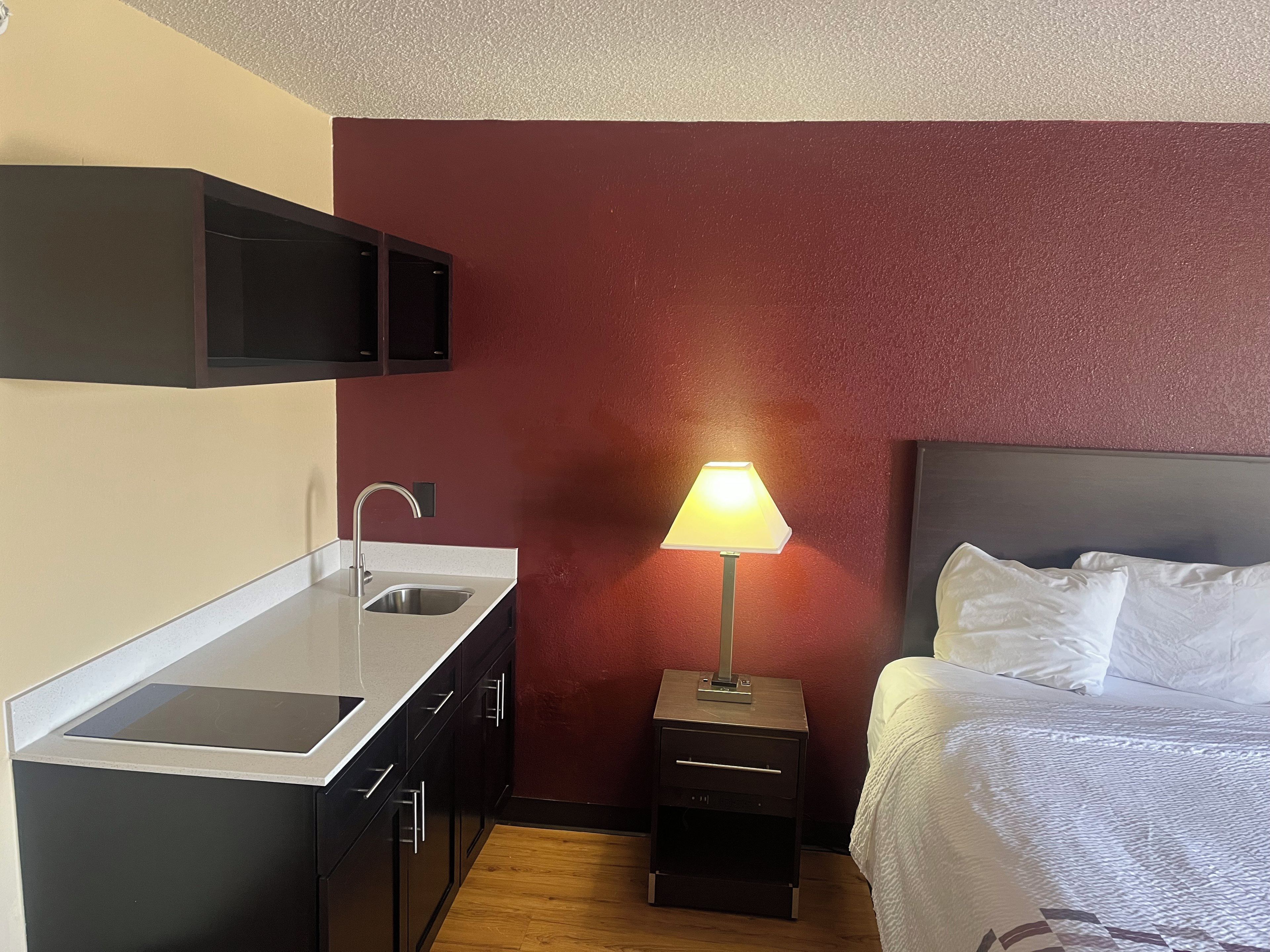 A compact hotel room with a kitchenette, a bedside table with a lamp, and a bed with white linens.