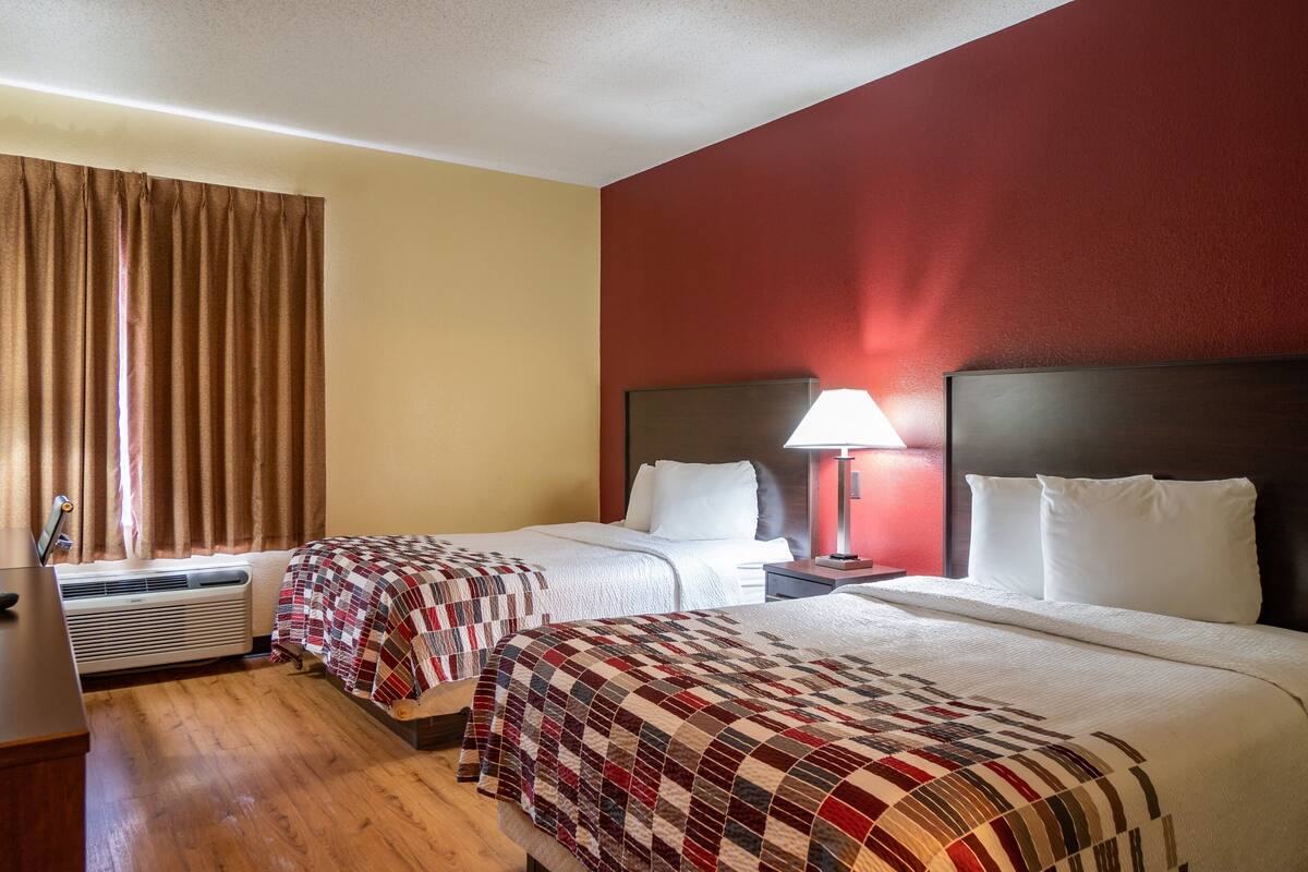 Een hotelkamer met twee bedden, een bureau, een lamp en een raam met gordijnen.