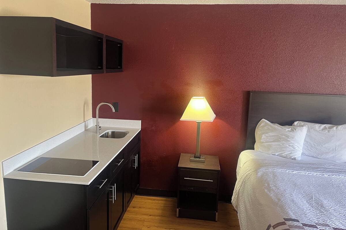 Een compacte hotelkamer met kitchenette, nachtkastje met lamp en bed met witte lakens.
