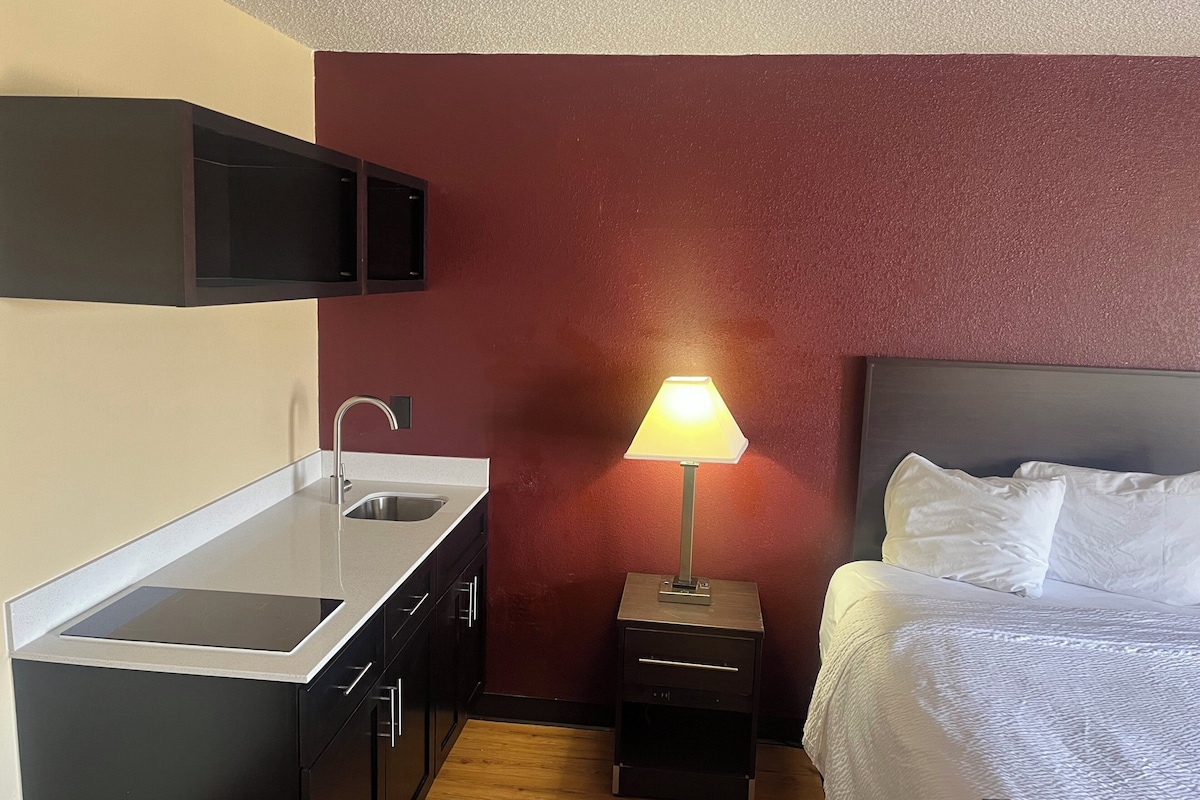 Ein kompaktes Hotelzimmer mit Küchenzeile, einem Nachttisch mit Lampe und einem Bett mit weißer Bettwäsche.