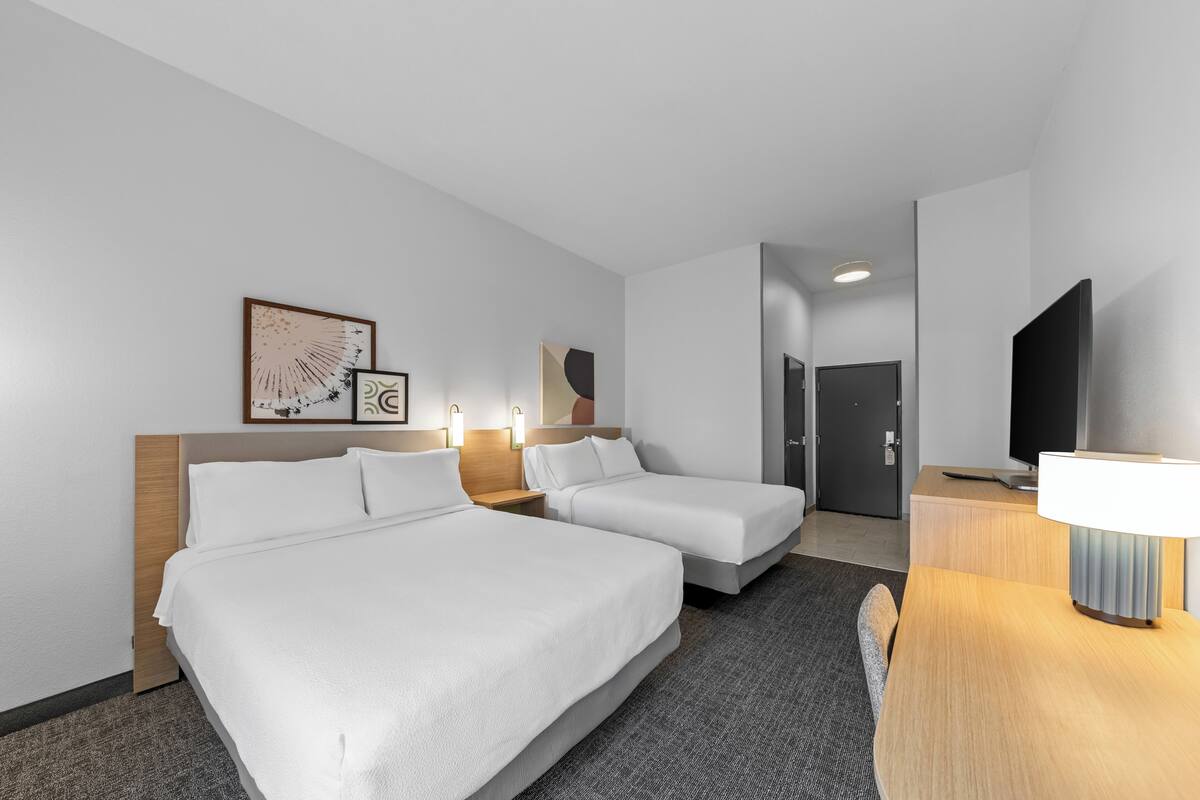 Una habitación de hotel con dos camas, un escritorio con lámpara, televisión y obras de arte en las paredes.