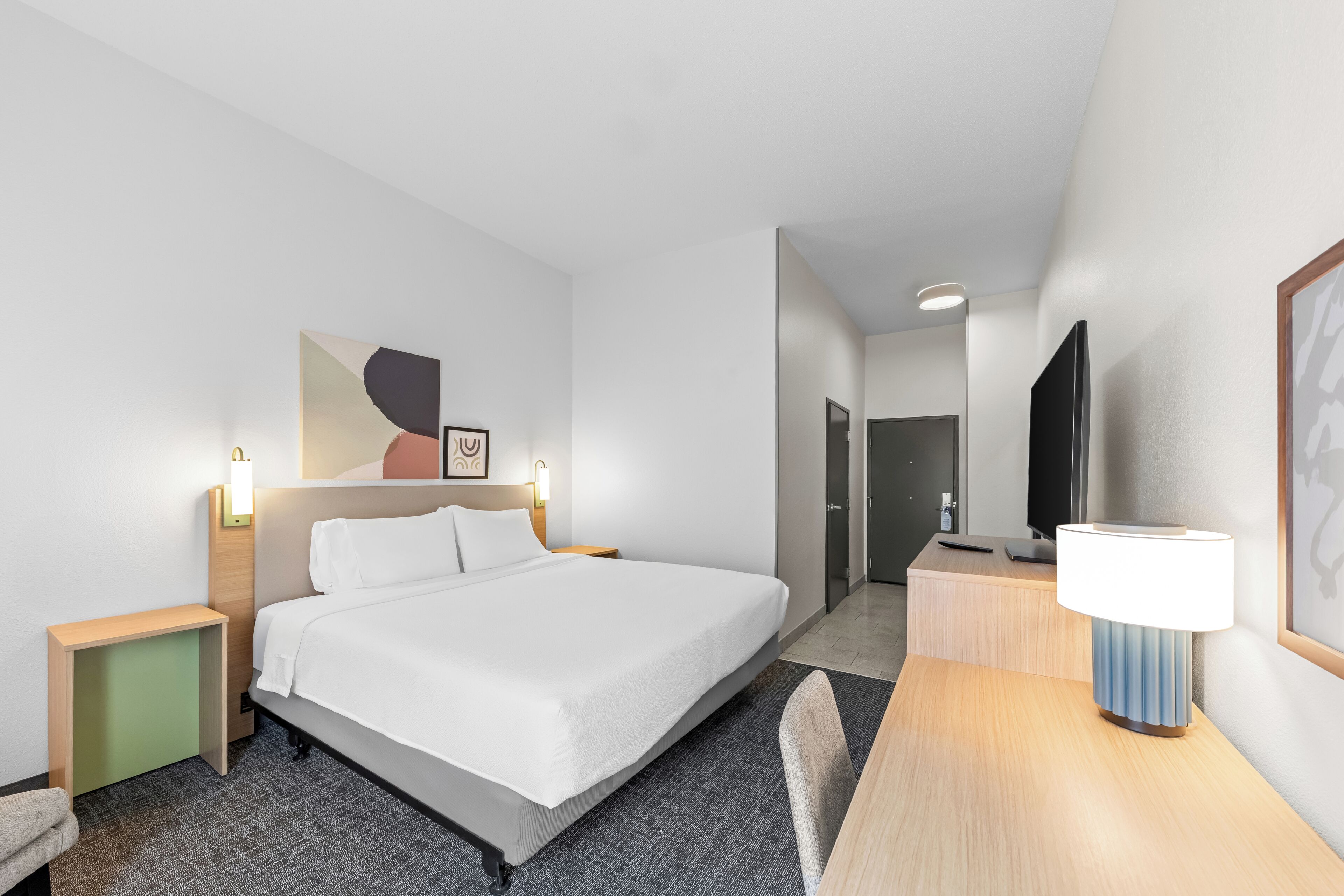 Habitación de hotel con cama, mesita de noche, lámpara, televisor y una obra de arte en la pared.