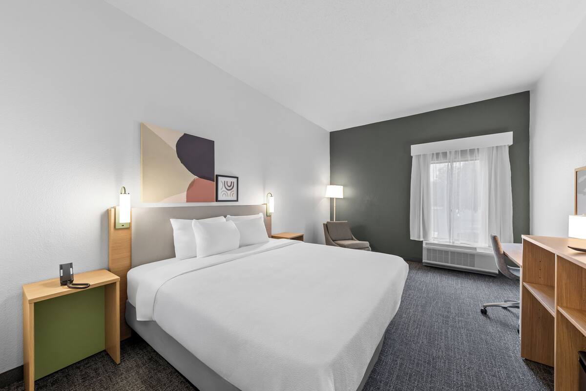 Una habitación de hotel con cama, mesitas de noche, escritorio, silla, ventana con cortinas y una obra de arte colgada en la pared.