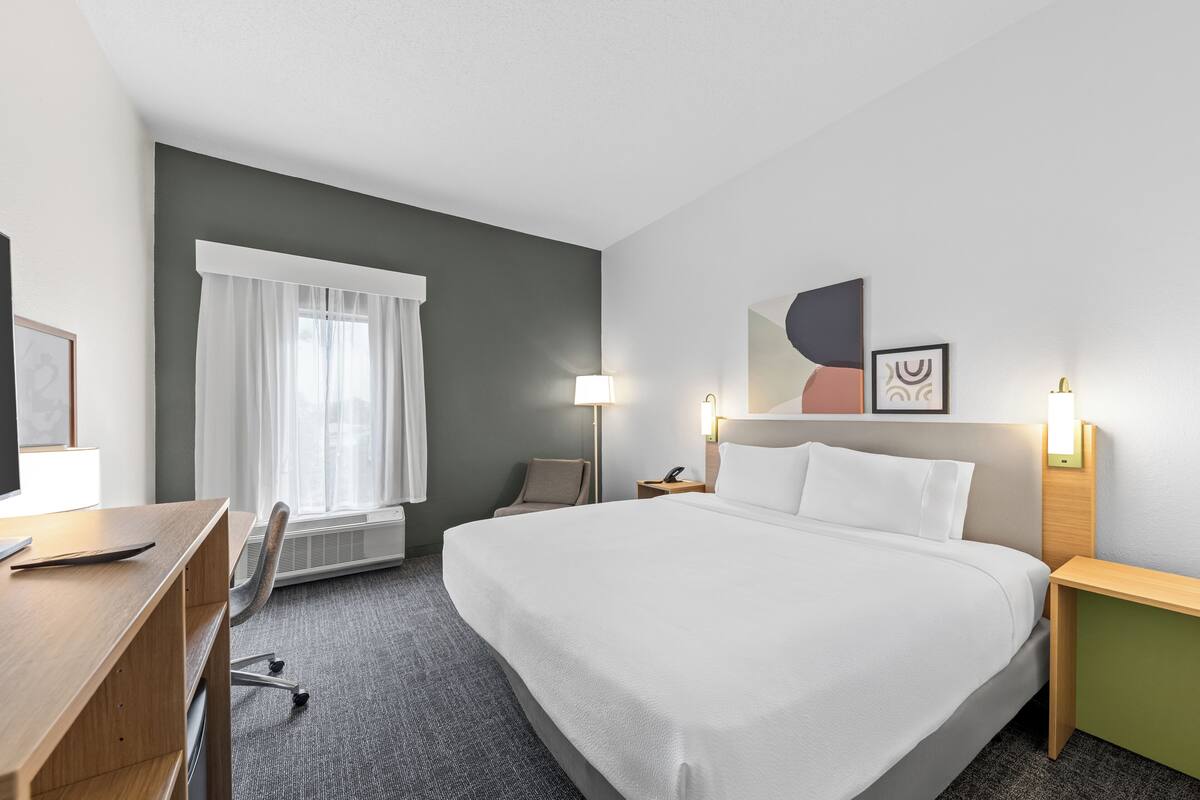 Una habitación de hotel con una cama grande, un escritorio, una silla, un ventanal con cortinas y lámparas de pared.