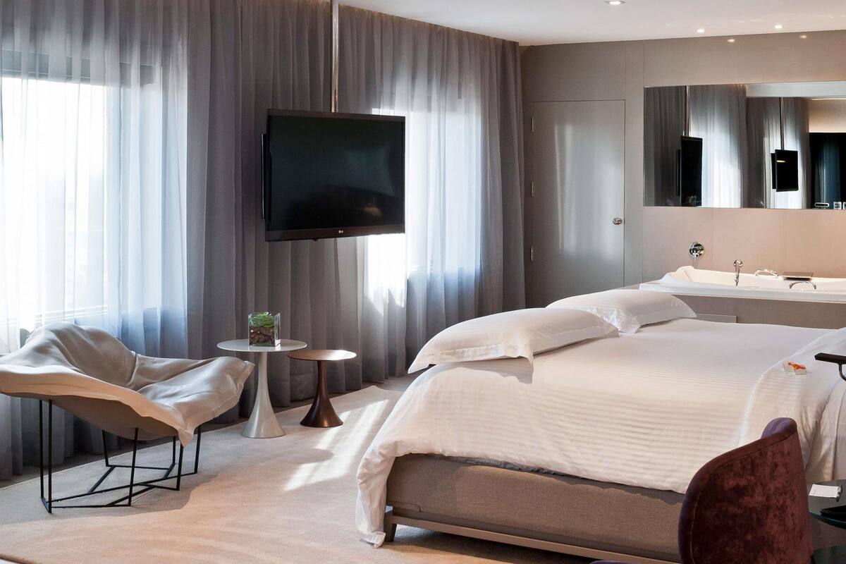 Een moderne hotelkamer met een groot bed, een flatscreen televisie aan de muur, een rond bijzettafeltje met een plant, een stoel en een grote spiegel boven het aanrecht.