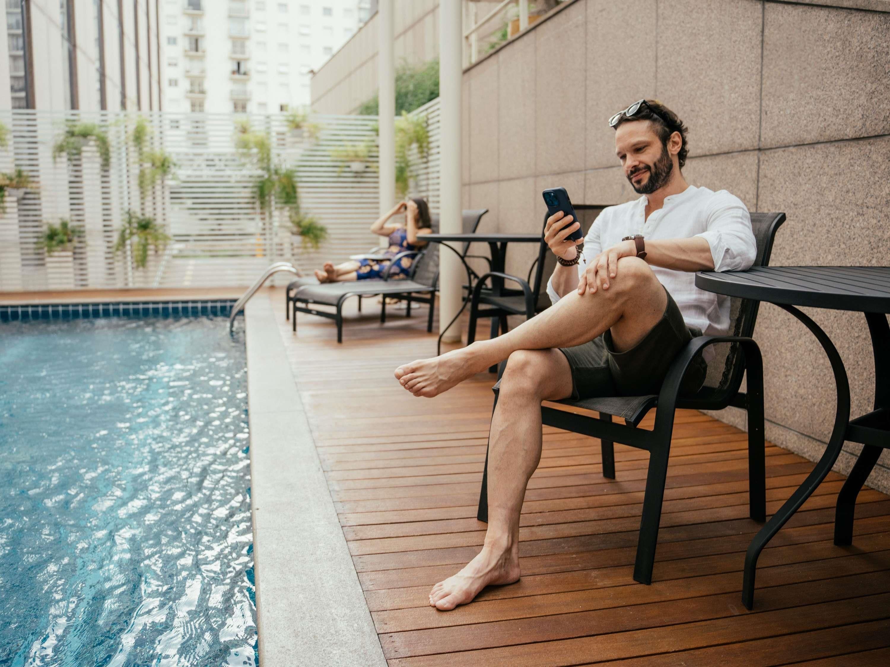 Um homem sentado à mesa junto à piscina, usando um smartphone.