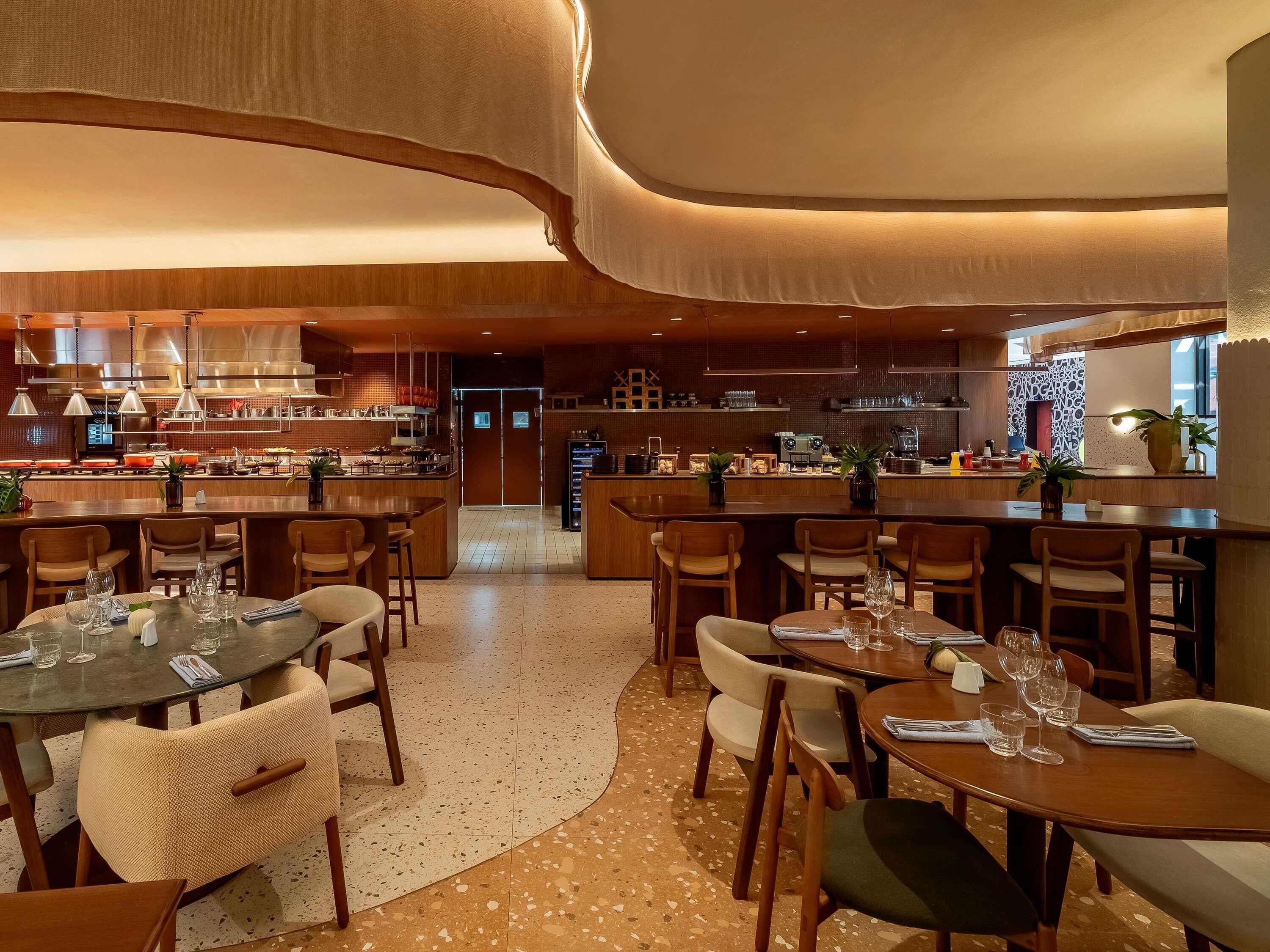 A modern restaurant with a bar area, wooden furniture, and a kitchen in the background.
