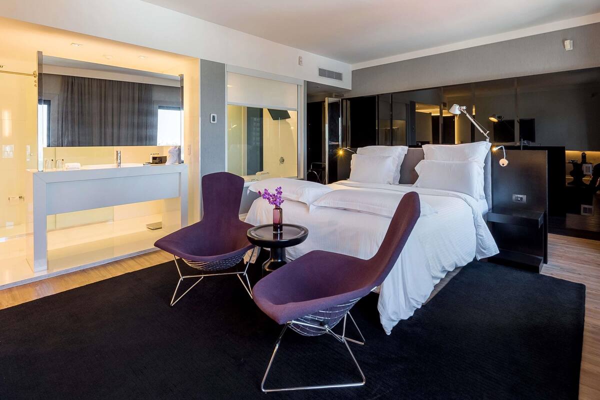 Een moderne hotelkamer met een groot bed, twee paarse stoelen, een tafeltje, een badkamer met een spiegel en een televisie.