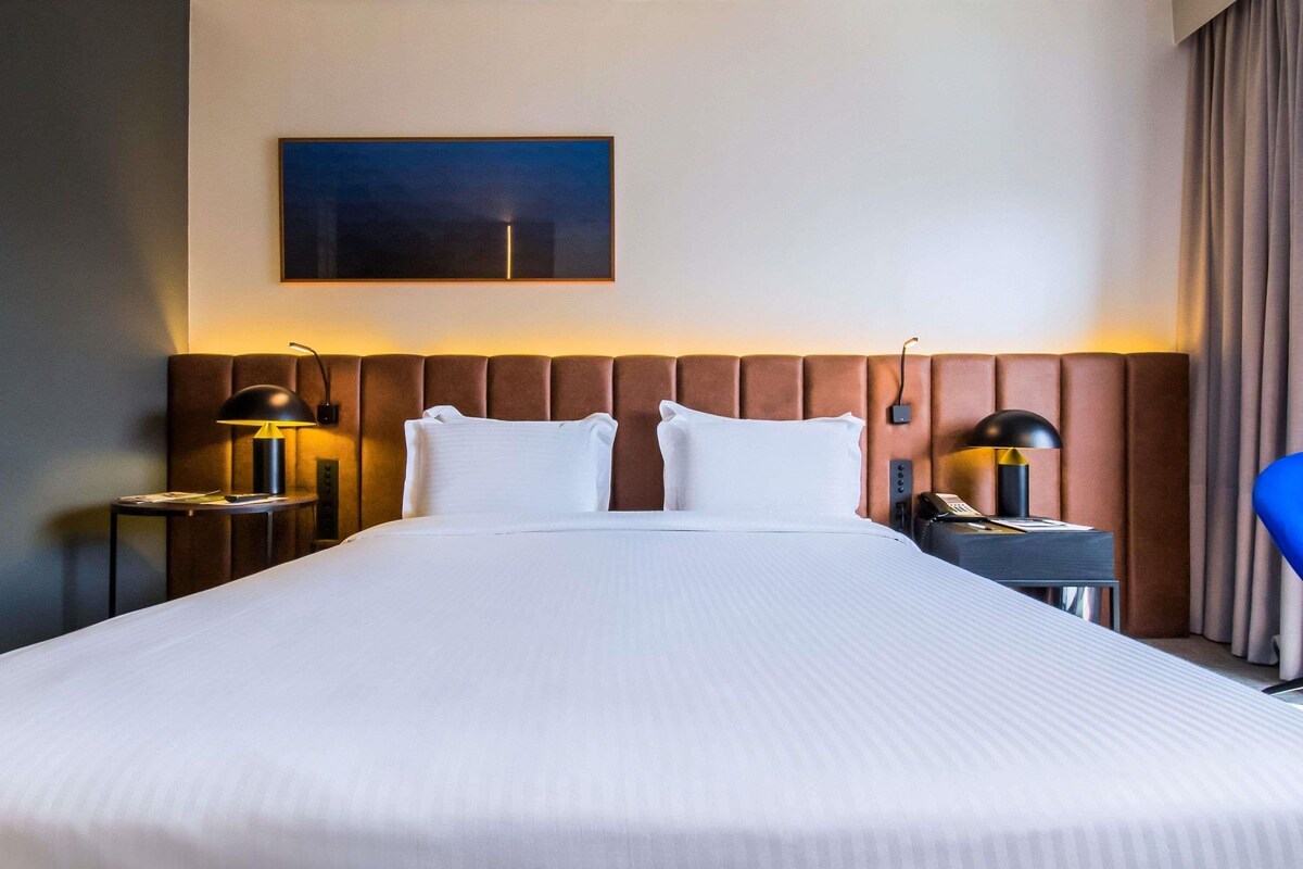 Een hotelkamer met een groot bed, twee nachtkastjes met lampen, een bijzettafeltje met telefoon, een blauwe stoel en een schilderij aan de muur.