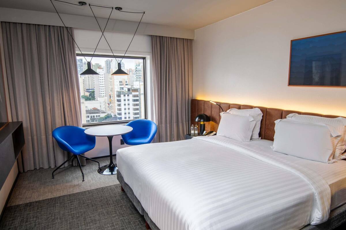 Een hotelkamer met een groot bed, twee blauwe stoelen, een klein tafeltje, een bureau, een lamp en uitzicht op de stad.