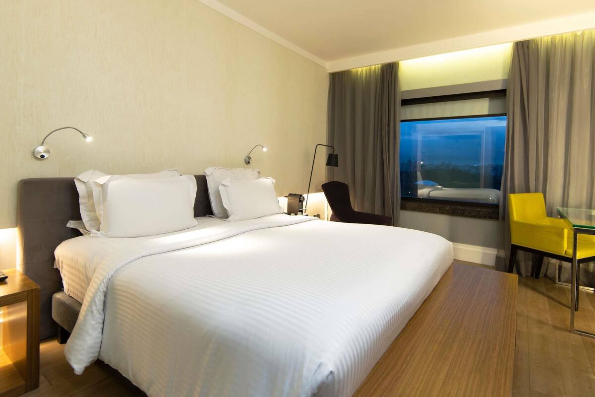Een hotelkamer met een groot bed, twee bedlampjes, een raam met gordijnen, een stoel en een klein tafeltje.