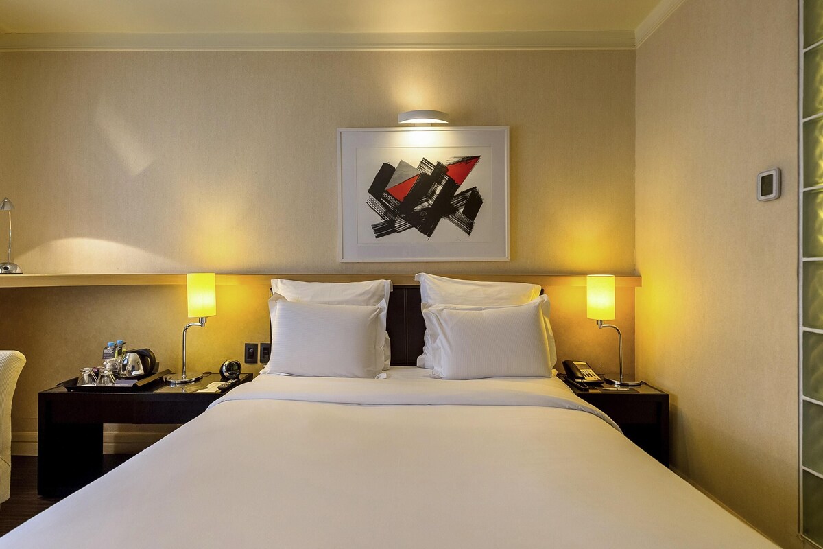 Une chambre d’hôtel avec un grand lit, deux tables de chevet, un bureau avec une lampe, une œuvre d’art abstraite encadrée accrochée au mur et une fenêtre avec des stores.