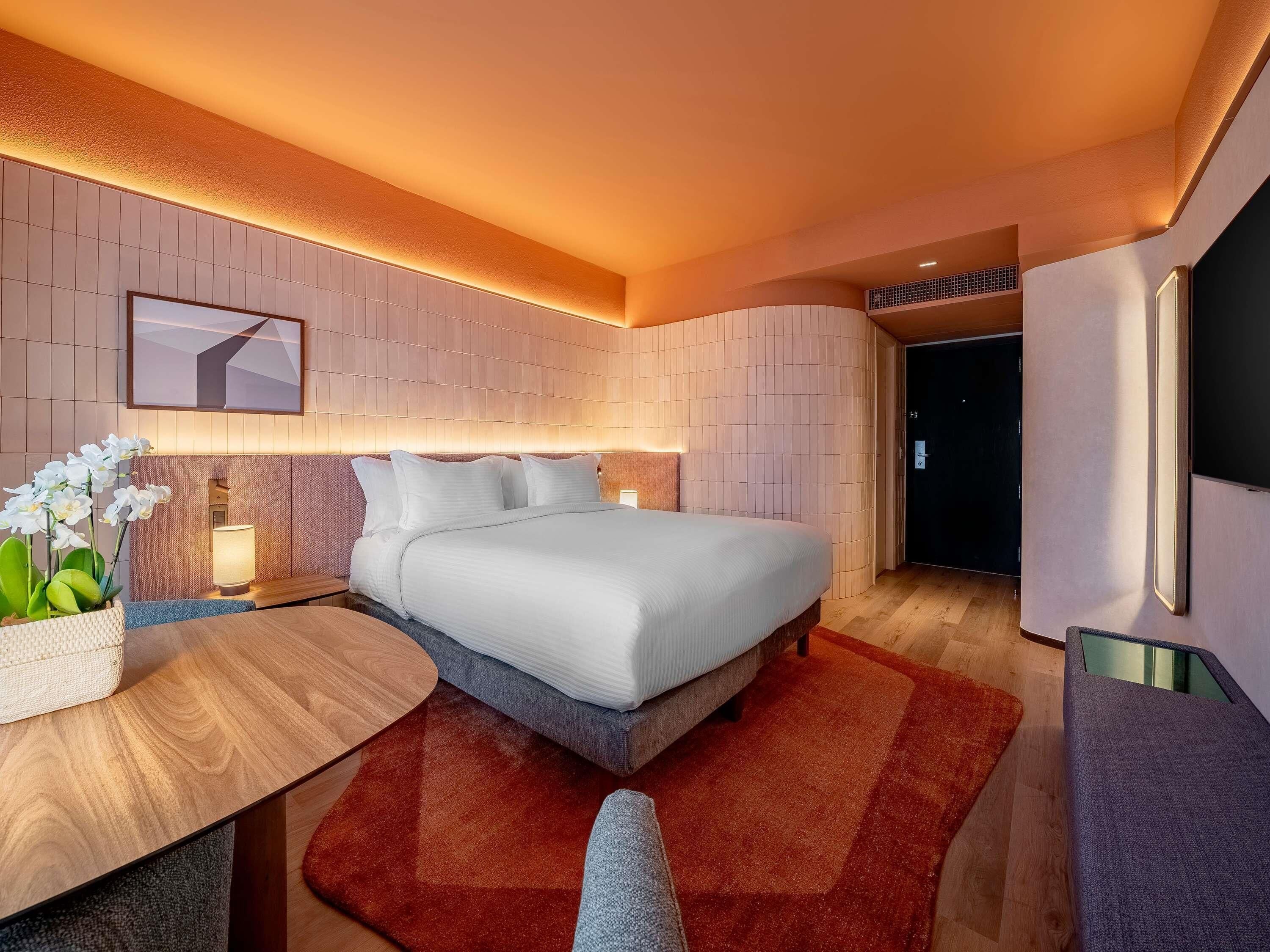 Ein modernes Hotelzimmer mit einem ordentlich bezogenen Bett, einem Nachttisch mit Lampe, einer kleinen Pflanze und einem roten Teppich.