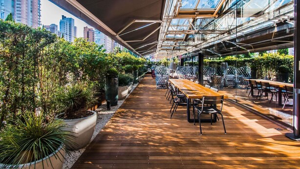 Un jardin intérieur avec des tables et des chaises en bois, entouré de plantes en pot et surmonté d’un plafond en verre.