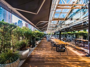 Un jardin intérieur avec des tables et des chaises en bois, entouré de plantes en pot et surmonté d’un plafond en verre.