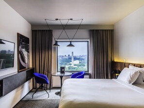 Ein Hotelzimmer mit einem Bett, einem Schreibtisch, zwei Stühlen und einem Blick auf die Stadt. An der Wand hängt ein Gemälde.
