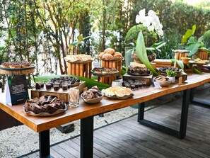 Una mesa de madera con diferentes alimentos y bebidas, rodeada de follaje y una terraza de madera.